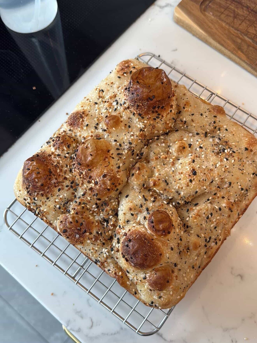 A freshly baked sourdough focaccia with black and white sesame seeds on a wire cooling rack, golden brown with deep dimples