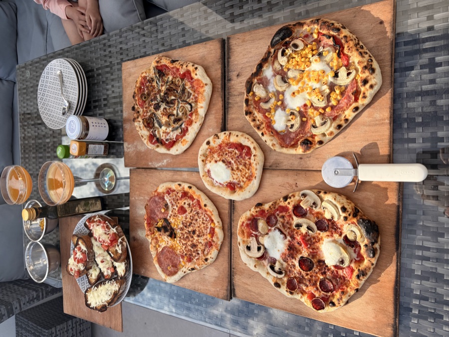 Four different sourdough pizzas laid out on wooden boards across an outdoor table with drinks, ready to eat