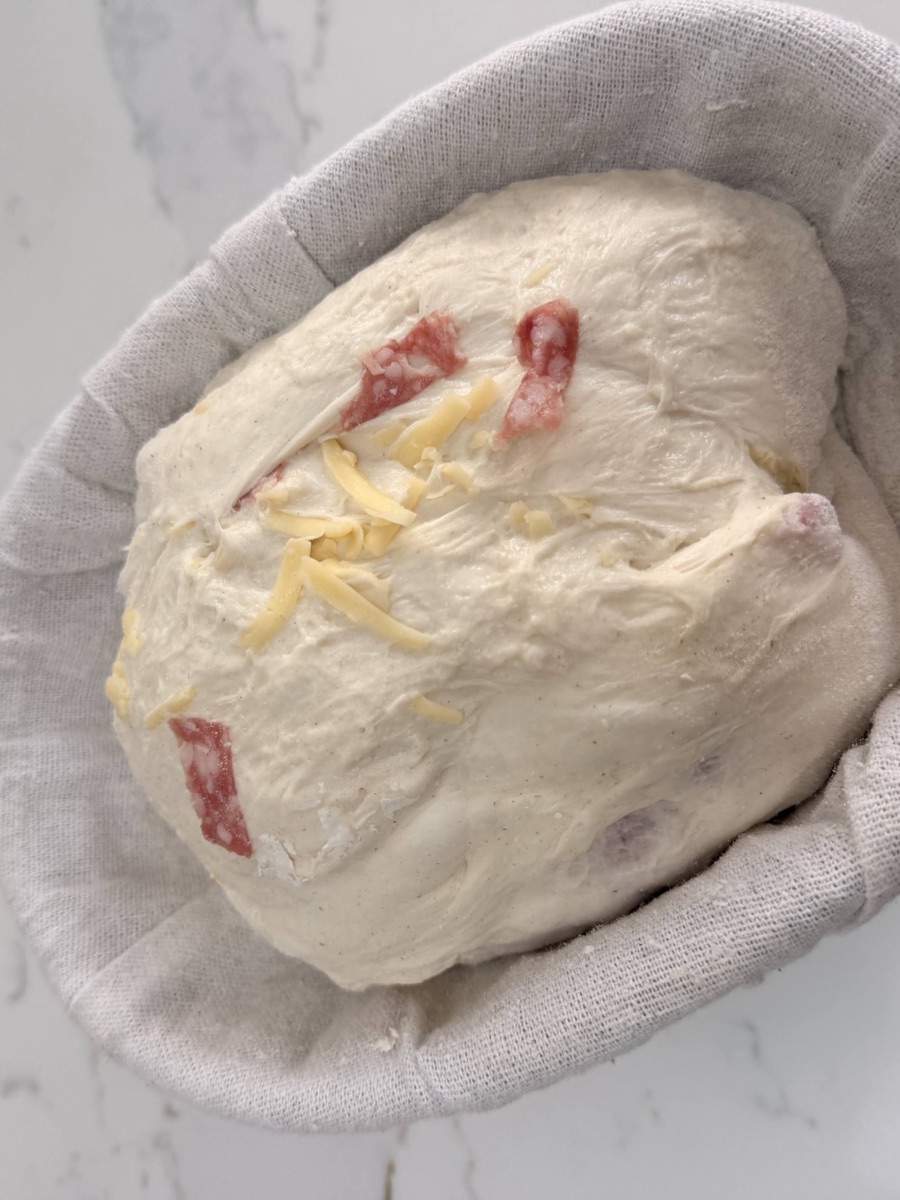Shaped salami and cheese sourdough sitting seam-side up in an oval banneton, with inclusions visible at the surface