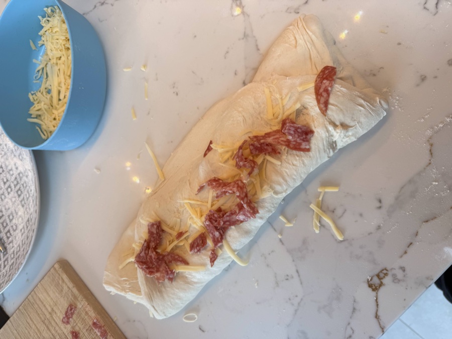 Sourdough dough pressed flat on a work surface with grated cheese and torn salami pieces scattered evenly across it