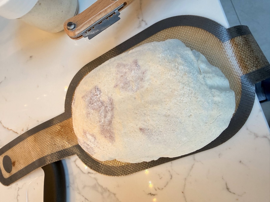 Salami and cheese sourdough turned out onto a baking mat, dusted with flour and ready to score, with a lame visible beside it